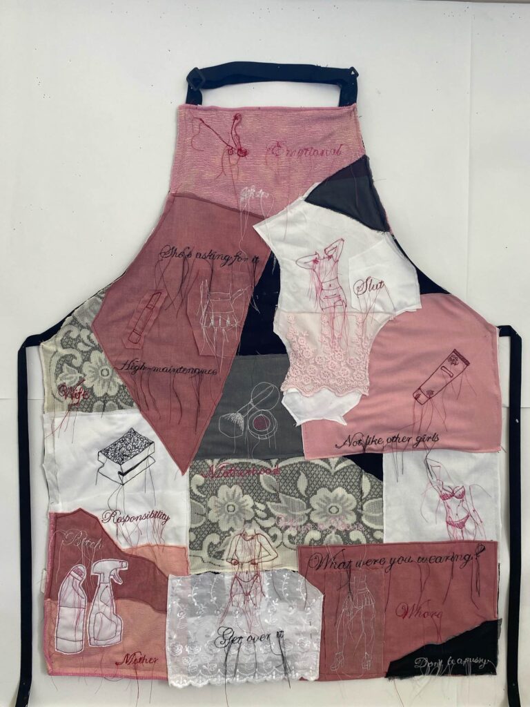 An patchwork apron made of various pink coloured fabric and lace. Embroidered various images associated to "women's duties" ie makeup, cleaning products, female bodies in the male gaze
Also embroidered are the following words: 
She's asking for it
Slut
High maintenance 
Wife
Motherhood
Responsibility
Bitch
Get over it 
Not like the other girls