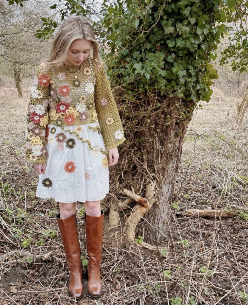 A young woman with blond hair standing next to a tree covered in ivy. She is wearing a crochet dress with colourful flowers covering the one sleeve and sprinkled across the garment 