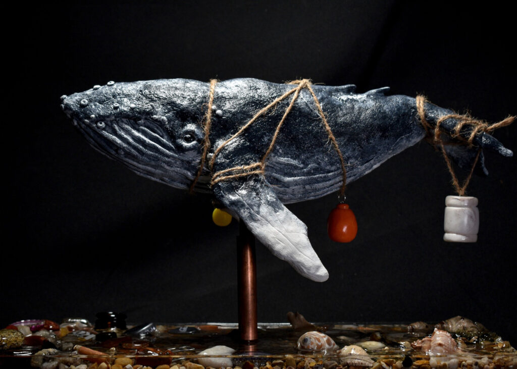 A sculpture of a whale with a rope encircling its body with various pieces of plastic objects hanging from it. 
Below the whale is shells, sand and various pieces of rubbish