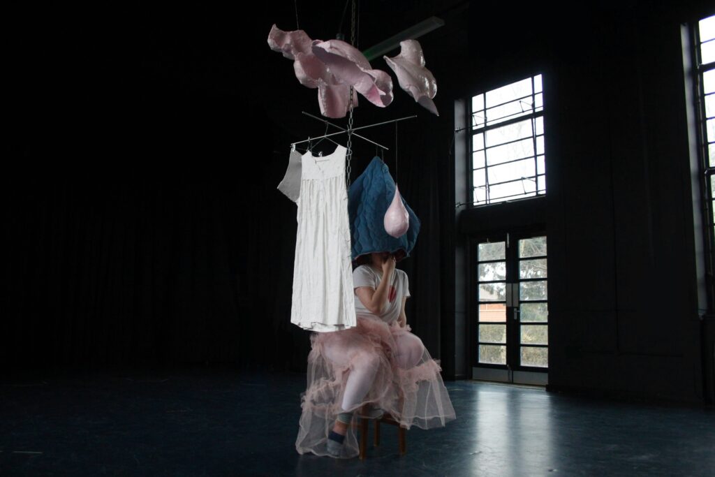 A large mobile sculpture with various forms in pink, with a white night dress and blue quilted fabric hanging from it. 
Below the fabric sits a young woman in a white t-shirt and pink ruffled skirt