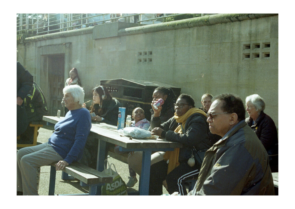 A street photograph of a diverse group sitting and standing around a 
a picnic table. Everyone is looking forward with the excepting of a young girl who is looking up at her mother.