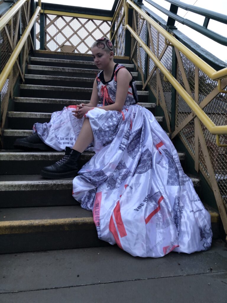 A young woman with a shaved head in a long white, black and red patterned dress sitting stairs. Dyed onto her hair are smiley faces