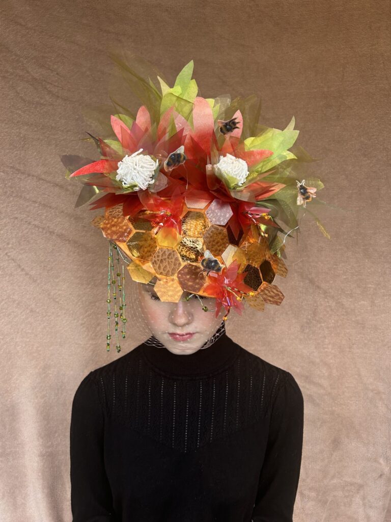 A young woman in a black top is wearing a hat, with a honeycomb pattern on it, it also has large flowers made of paper, fabric and beads. Hovering over the flowers on wire are bees. 