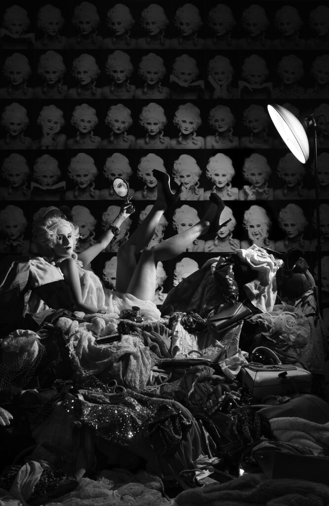 A young woman in a white historical wig with pearls is sitting on a pile of clothes and accessories she holds a mirror in front of her but she is looking at the viewer. In the background the wallpaper is portrait photographs of herself in different poses.