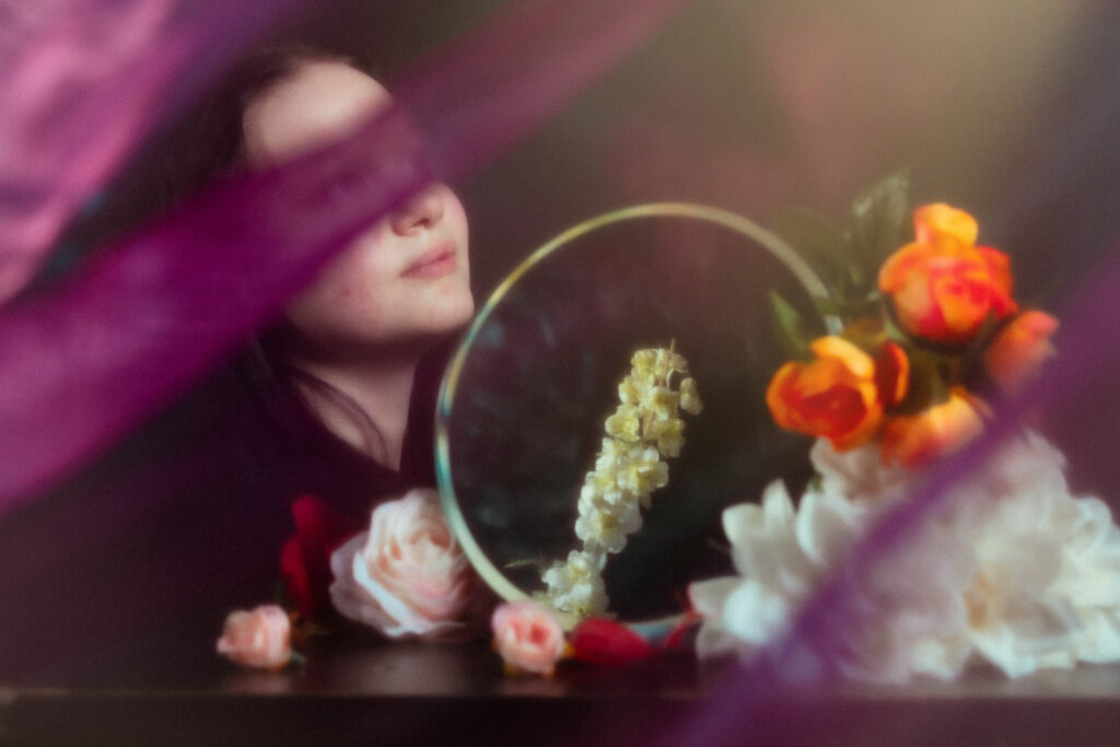 A dreamy digital photograph capturing a woman's serene face and vibrant flowers, partially obscured by soft purple veils, taken with a Nikon Z-50 mirrorless camera.