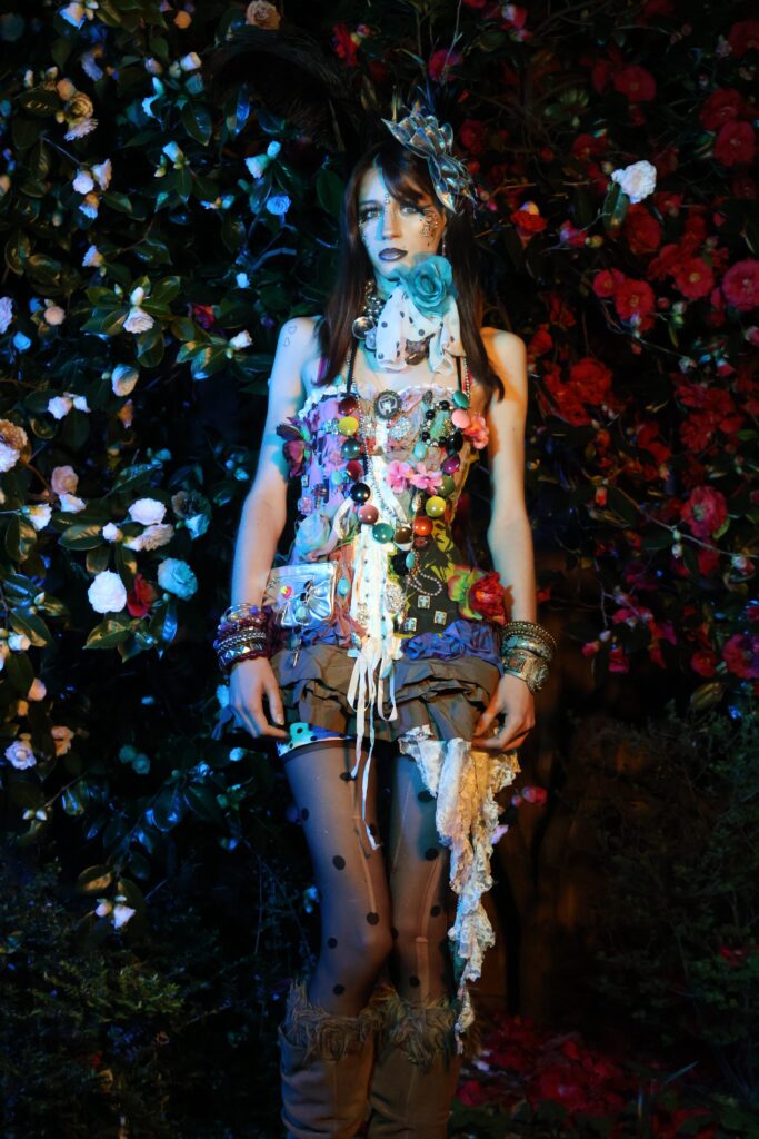 A fashion photograph of a model wearing a highly embellished garment made with mixed textures, beads, and flowers, posed against a background of flowers.