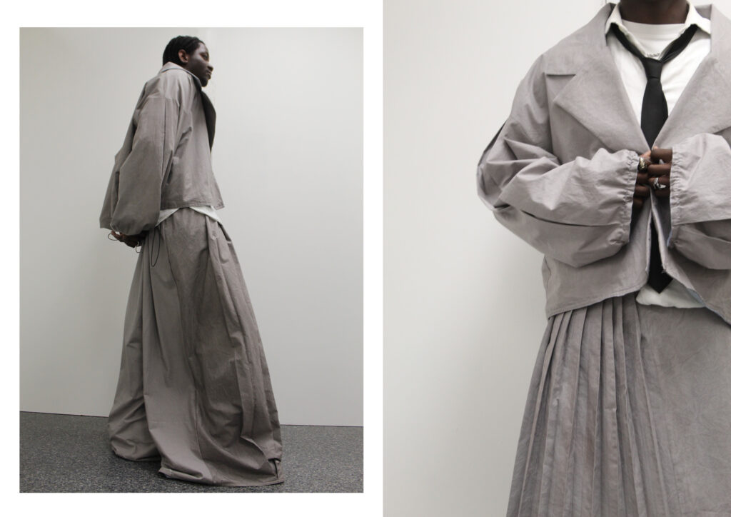 A fashion photograph of a model wearing wide-cut grey clothing, including a pleated skirt and an oversized jacket, styled with a white shirt and black tie against a plain backdrop.