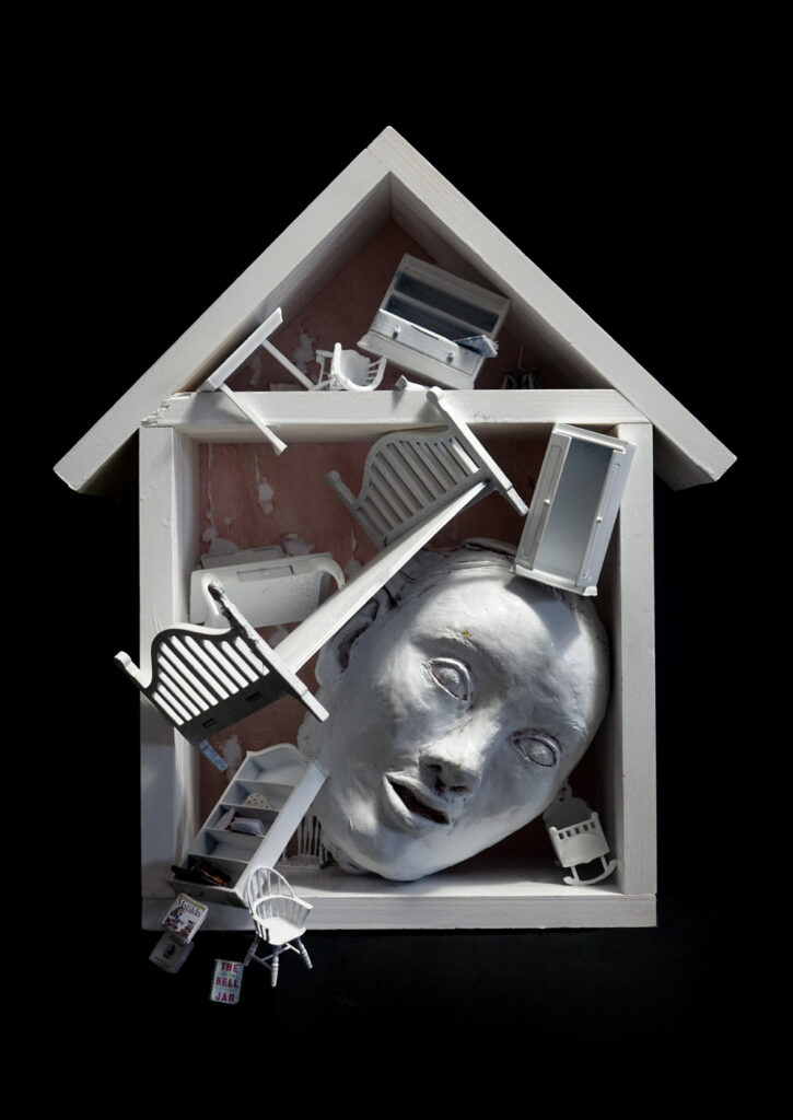 A mixed-media sculpture inside a house-shaped frame, featuring a large sculpted head surrounded by miniature white furniture, books, and objects.