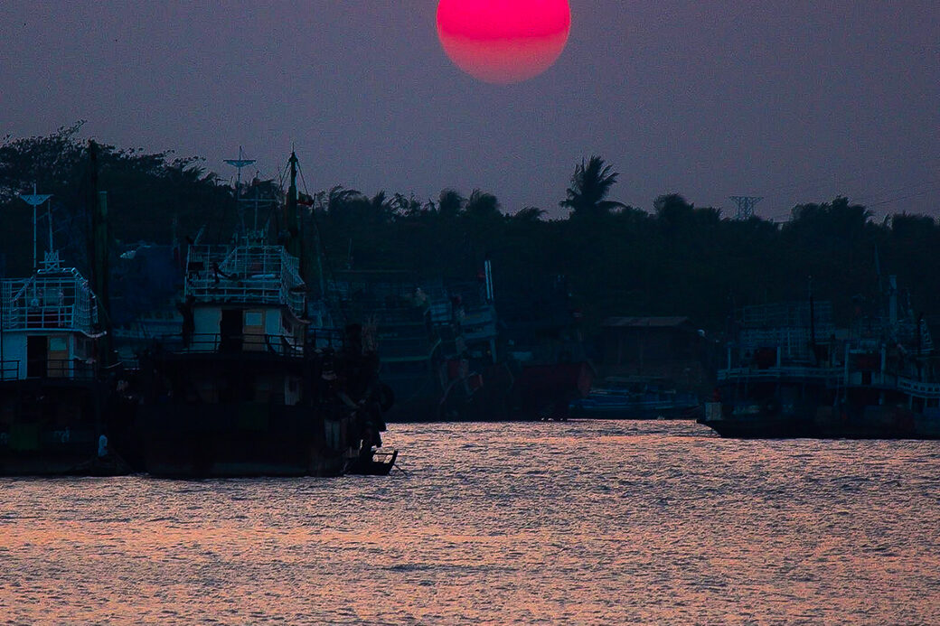 A sunset photograph with a vivid red sun low in the sky, silhouetting boats and figures on the water.