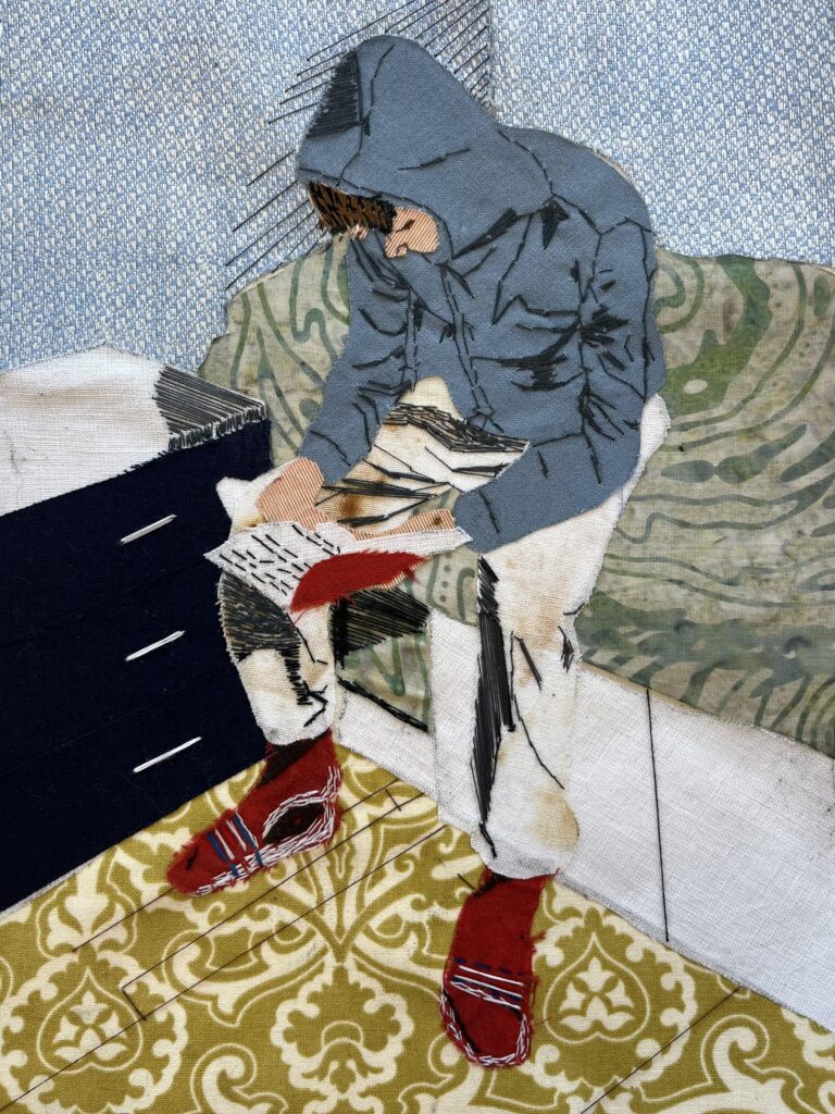 A textile artwork showing a young person in a hoodie reading, created using layered fabric and embroidery.