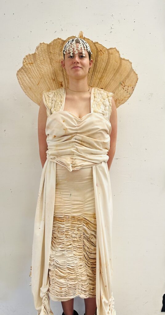 A person wearing a ruched cream garment with a sculptural shell-like collar and headpiece, referencing sea forms and protection.