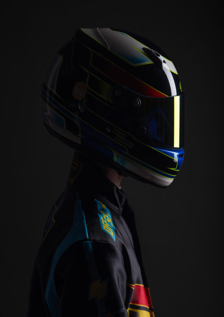 A studio photograph showing a person in racing gear and helmet, shot in profile against a dark background with focussed lighting.