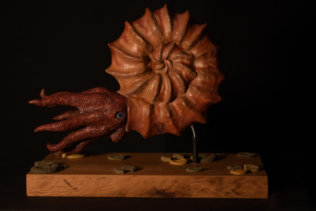 A sculpture of an ammonite with octopus-like limbs, displayed on a wooden base with small scattered fossils.