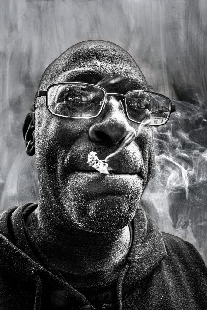A high-contrast black-and-white portrait of a man with glasses smoking, captured with sharp texture and detail.