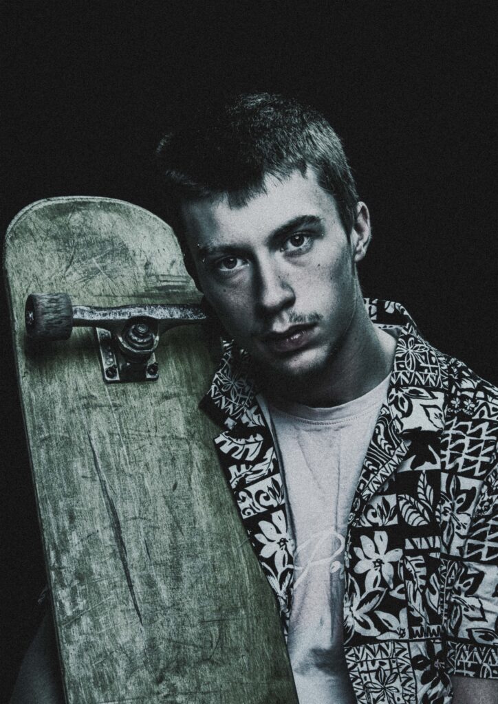 A photograph of a young man in patterned shirt holding a skateboard, against a dark backdrop.