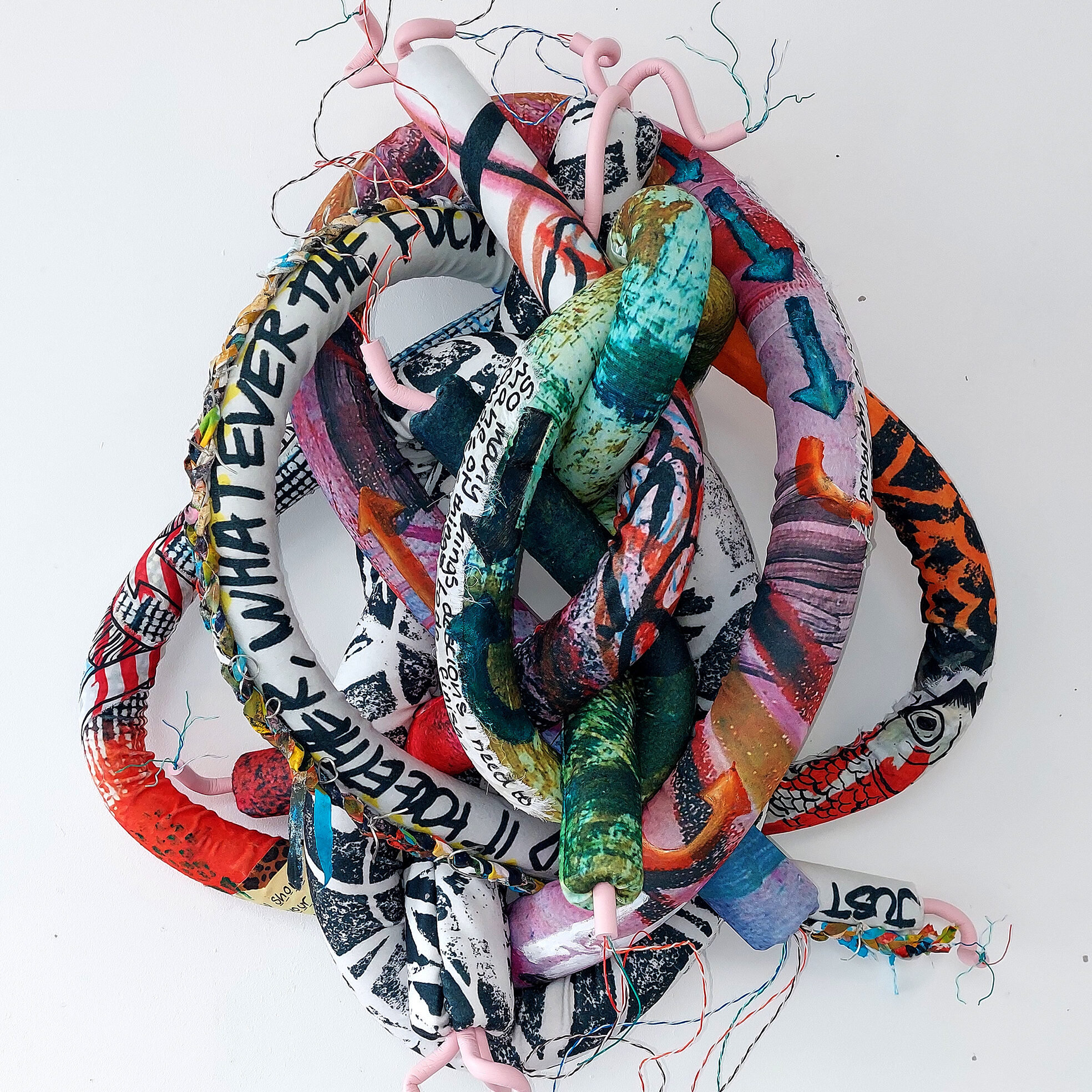 A sculptural knot made from colourful, printed tubes and wires, suspended against a white wall.